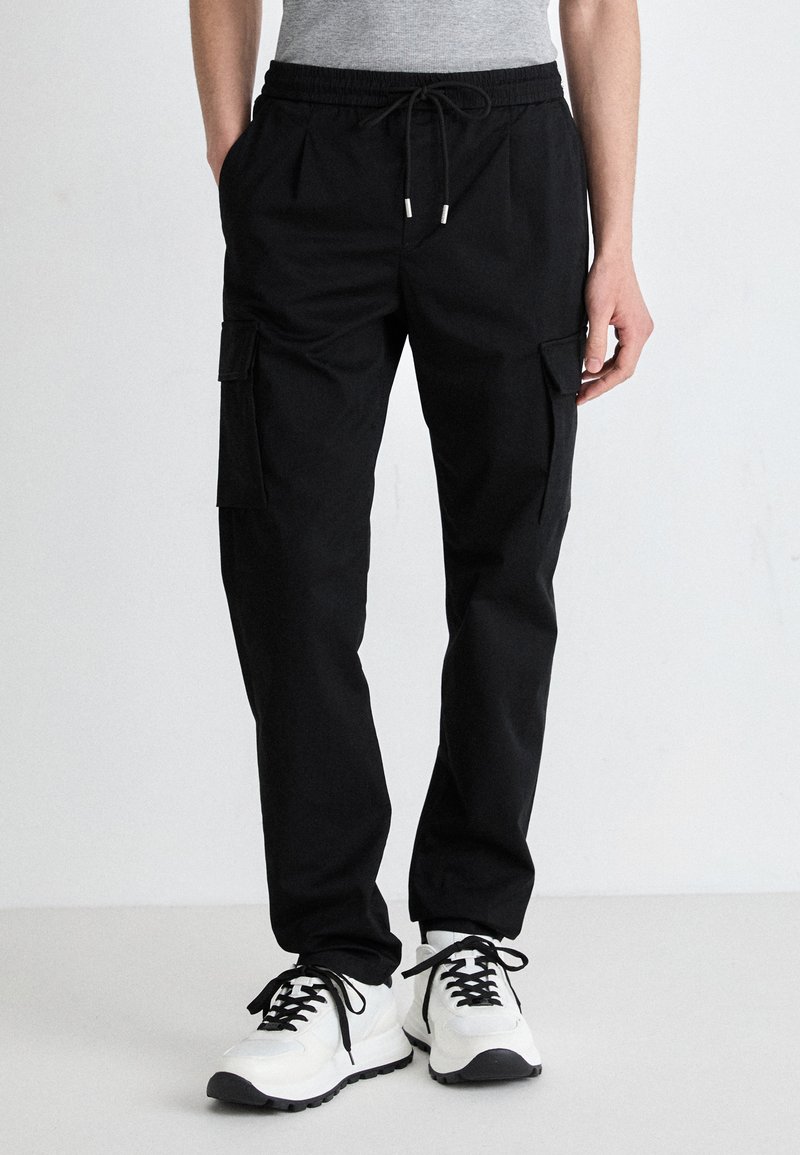 Schwarze Cargo-Hosen mit einem Kordelzugbund, mehreren Seitentaschen und einer lockeren Passform, kombiniert mit weißen Sneakers und einem grauen Oberteil.
