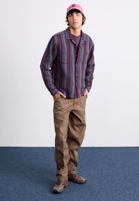Multicolored button-up shirt with vertical stripes in purple, blue, and green, paired with beige pants and brown shoes, complemented by a pink cap.