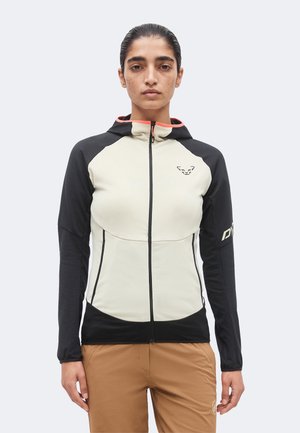 Persoană care poartă o jachetă cu fermoar neagră și bej cu glugă și pantaloni maro, stând într-un fundal simplu și deschis.