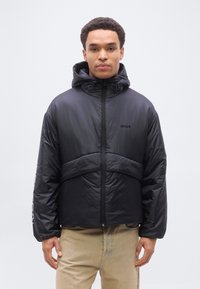 Άνδρας που φοράει μαύρο επενδυμένο μπουφάν με κουκούλα και λογότυπο «HUGO» στο στήθος και μπεζ παντελόνι, μπροστά σε απλό φόντο.