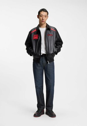 Jonge man die een zwart leren jack draagt met rode logo's, een wit t-shirt, donkere spijkerbroek en zwarte sneakers met rode accenten, staand met zijn handen in zijn zakken.