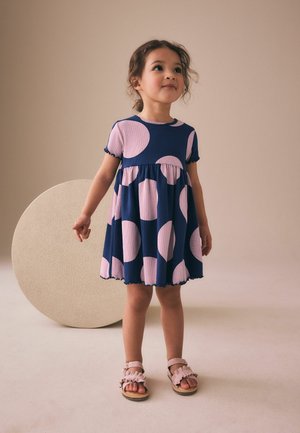Young girl wearing a navy blue dress with large pink circles and pink sandals, standing in front of a textured beige circular backdrop.