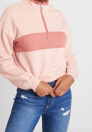 Kvinde iført en lyserød langærmet sweatshirt med en mørkere pink stribe over brystet og blå slidte jeans.