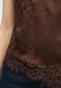 Αμάνικη κορυφή από καφέ σατέν με δαντέλα με λουλούδια, φορεμένη με σκούρο μπλε τζιν, που δείχνει τον κορμό και το μπράτσο του φορέα.