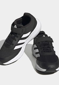 Baskets de sport noires avec une tige en mesh texturé, présentant des accents en trois bandes blanches, un design à lacets et à bandage Velcro, et une semelle rembourrée.
