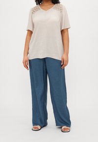 Donna che indossa una blusa beige a maniche corte con dettagli in pizzo e pantaloni larghi blu, in piedi con sandali bianchi su uno sfondo bianco.