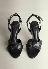 Schwarze Leder-High-Heelsandalen mit einem geknoteten Schleifendetail, gestifteten Akzenten und dünnen Knöchelriemen. Quadratische Zehenpartie und glatte Textur.