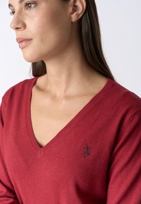 Maglione rosso a scollo a V in tessuto lavorato a maglia, con bordi a costine e un piccolo logo ricamato sul lato sinistro del petto.