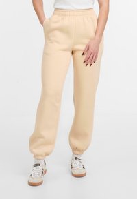 Beige Sweatpants aus weichem Material mit elastischem Bund und schmalen Bündchen. Kombiniert mit weißen Sneakers und sichtbaren Socken.