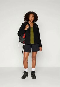 Kvinne med krøllete hår iført en svart jakke, olivengrønn topp, svarte shorts, tursko, hvite sokker og bærende en svart og rød ryggsekk.