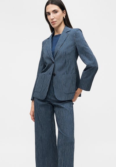 Γυναίκα με μπλε σκούρο σακάκι με ρίγες και ταιριαστό παντελόνι με φαρδιά μπατζάκια, στέκεται με το ένα χέρι στη μέση μπροστά σε μονόχρωμο φόντο.