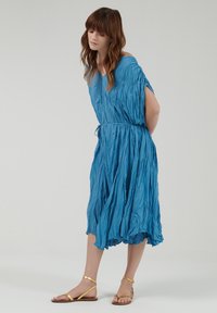 Blauwe, gekreukte stoffen jurk met een V-hals en korte mouwen; verzamelde taille met een trekkoord; enkellange zoom. Draagt bijpassende gouden sandalen.