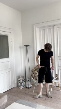 Camiseta negra ajustada, pantalones cortos negros y zapatillas color marrón claro. De pie en una habitación minimalista con suelos de madera y farolillos decorativos.