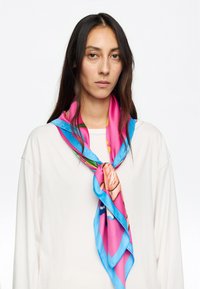 Foulard di seta con colori vivaci di rosa e blu, decorato con motivi floreali. Texture liscia, annodato attorno al collo in uno stile stratificato.
