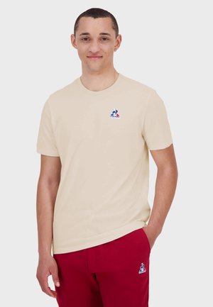 T-shirt crème à manches courtes en coton, avec un col rond et un petit logo bleu et rouge sur la poitrine gauche.