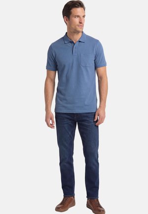 Mann steht und trägt ein blaues kurzärmeliges Poloshirt mit Brusttasche, dunkelblaue Jeans und braune Ledersneaker vor weißem Hintergrund.