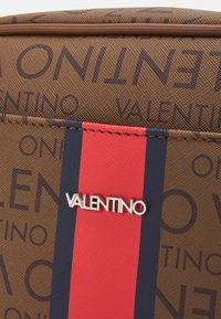 Brown textured fabric with repeated "Valentino" text, featuring a vertical red and navy blue stripe and a metallic Valentino logo.