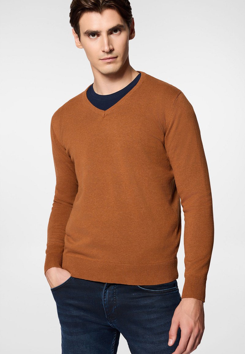 Sweter w kolorze rdzy z dekoltem w serek, wykonany z gładkiego dzianego materiału, z prążkowanymi mankietami i dołem, noszony na granatowej koszulce z okrągłym dekoltem. Dopasowany krój.