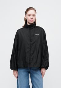 Kvinde med lys hud og rødt hår iført en løs sort lynlåsjakke med høj krave og blå jeans mod en ensfarvet hvid baggrund.