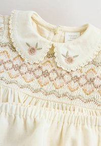 Robe pour bébé de couleur crème avec un col festonné et une broderie florale délicate dans des teintes de rose doux et de vert, agrémentée de détails en smocks.