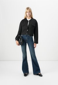 Svart avkortet jakke med knappelukking, to brystlommer, sammen med utsvingte blå denimjeans. Modellen holder en liten svart håndveske.