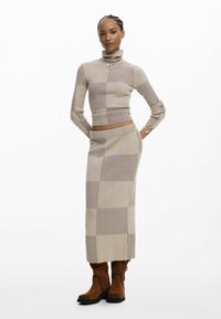 Beżowy dzianinowy komplet dwuczęściowy z golfem w pionowe paski i długą, dopasowaną spódnicą. Uzupełniony srebrnym naszyjnikiem w kształcie serca.