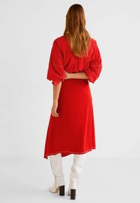 Robe rouge avec manches bouffantes, longueur mi-mollet. Présente une texture lisse et un contraste de passepoil blanc à la taille et à l'ourlet. Associée à des bottes blanches.
