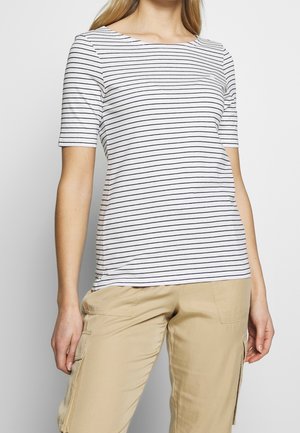 Frau trägt ein weißes kurzärmeliges Hemd mit dünnen schwarzen horizontalen Streifen und beigefarbenen Cargohosen vor einem schlichten Hintergrund.