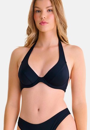 Jonge vrouw draagt een zwarte halterbikinitop en bijpassende broekjes tegen een effen lichte achtergrond.