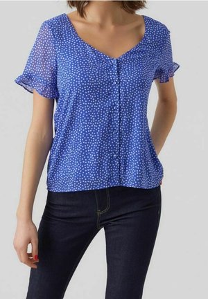 Femme portant un chemisier bleu à manches courtes avec des pois blancs et des bords froncés, associé à un jean bleu foncé.