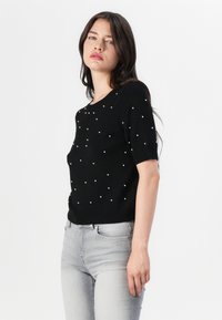 Pull noir à manches courtes avec un col rond, orné de décorations blanches dispersées, fabriqué dans un matériau tricoté doux.