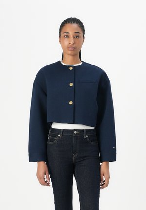 Kuttet marineblå jakke med rund krage, gullknappelukking, enkelt brystlomme og strukturerte ermer, kombinert med mørke jeans.