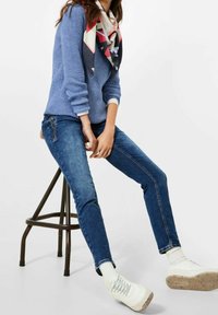 Blauwe gebreide trui, donkerblauwe jeans met een zijrits, witte sneakers met gestructureerde zolen en een kleurrijke, patroon gesjaald.