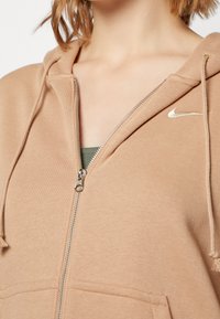 Kvinna i en delvis dragkedjad tan Nike-hoodie som visar en grön topp under, med fokus på övre bröst- och nackområde.