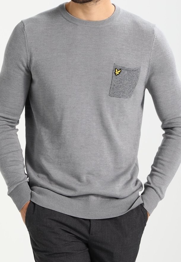 Homme portant un pull gris clair avec une poche poitrine gris foncé ornée d'un petit logo d'oiseau jaune, associé à un pantalon gris foncé.