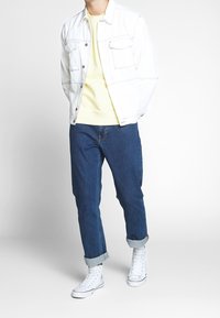 Giacca di denim bianco sopra un maglione giallo chiaro, abbinata a jeans blu scuro con risvolti e scarpe da ginnastica bianche. Design strutturato con tasche.