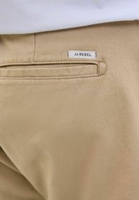 Poche arrière d'un pantalon beige avec étiquette JJ REBEL, porté avec un haut blanc.