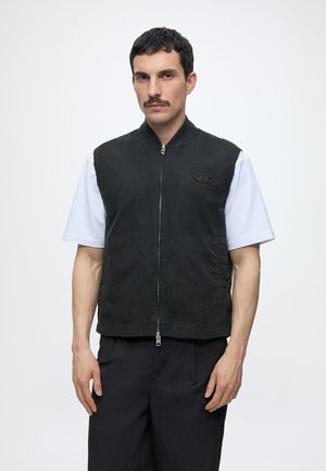 Mann mit dunklen Haaren und Schnurrbart, der eine schwarze ärmellose Reißverschlussweste über einem weißen kurzärmeligen Hemd vor einem schlichten weißen Hintergrund trägt.