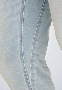 Približnji posnetek lahkih modrih jeansov, ki prikazujejo stranski šiv in teksturirano tkanino z nežnim bledenjem.