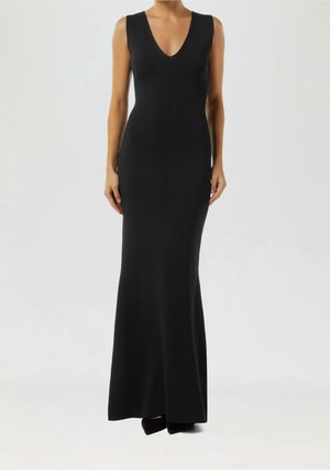 Kvinde iført en ærmeløs, tætsiddende sort maxikjole med dyb V-udskæring, stående foran en enkel lys baggrund.