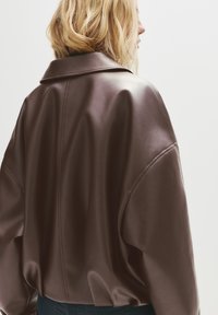 Giacca in finto pelle marrone con un ampio colletto, vestibilità over-size e dettagli cuciti lungo la schiena. Tessuto liscio con un design drappeggiato.