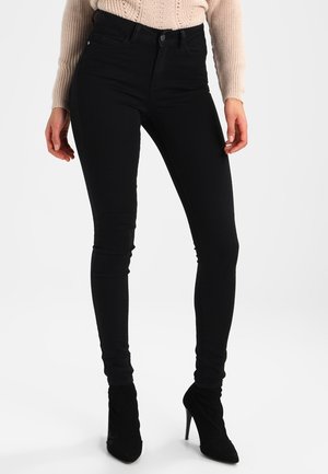 Jeans ajustados negros hechos de algodón elástico; diseño de cintura alta con una textura suave y detalles de costura sutiles. Combinados con tacones negros.