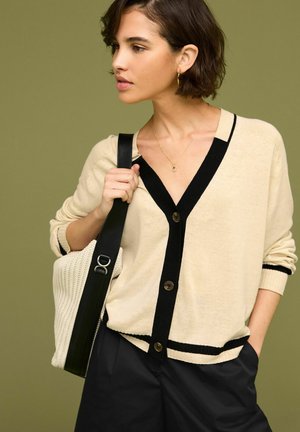 Beige cardigan met zwarte afwerkingen en knopen, met een diepe V-hals. Draagt het samen met een zwarte wijde pantalon, gecombineerd met een lichtgeweven witte tas.