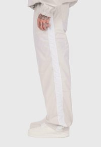 Cilvēks, kurš valkā gaiši bēšas bikses ar baltu sānu svītru un baltas kedas, rādot tetovētu roku un daļēju piedurkni.