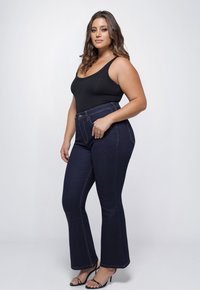 Donkerblauwe high-waisted flared jeans met contrasterende stiksels. Model draagt een zwart tanktop en zwarte sandaaltjes met straps.