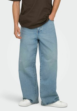 Muž nosiaci voľné svetlomodré rifle, biele tenisky a tmavohnedé tričko, s jednou rukou v vrecku, na jednofarebnom pozadí.