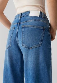 Vaqueros de mezclilla azul claro con cintura alta y diseño de pierna ancha. Incluyen dos bolsillos traseros y una etiqueta blanca en la pretina.