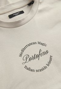 Beige Baumwoll-T-Shirt mit einem runden Grafikdruck, auf dem der Text "Mediterranean Magic Portofino Italian seaside leisure" in Schwarz steht.