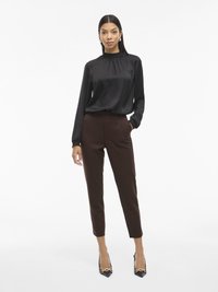 Melnas satīna blūze ar augstu apkakli un garām piedurknēm, kopā ar pielāgotām brūnām biksēm un melniem kurpēm ar norādītu degunu, kas rotātas ar zelta akcentiem.