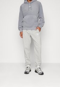 Felpa grigia con tasca frontale e logo, abbinata a pantaloni tuta grigi chiari con un piccolo logo. Calzature grigie e nere con lacci.